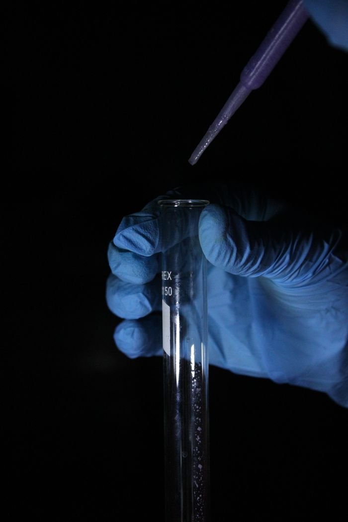 who-we-are Blue-gloved hand holds a test tube while a pipette dispenses liquid in a lab.